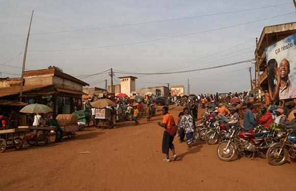 A bustling street scene with people and motorcycles, various stalls and shops, and buildings under a clear sky.