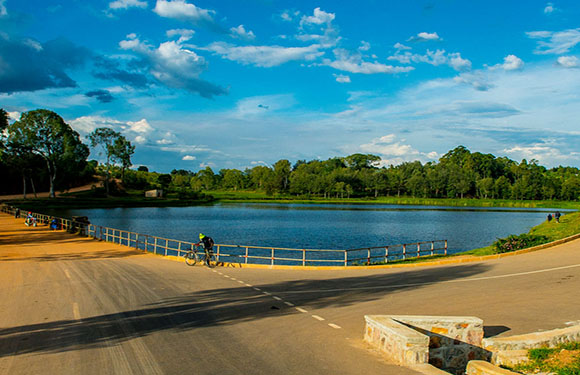 A serene lake scene with a blue sky, fluffy clouds, a paved path with railing, green trees, and shadows suggesting morning or afternoon.