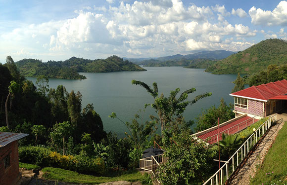 A scenic view of a lake surrounded by green hills under a partly cloudy sky, with a red-roofed building on the right.