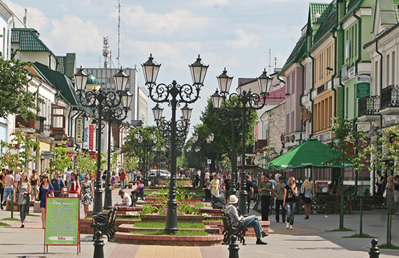 A busy pedestrian avenue with trees, benches, and European-style buildings under a partly cloudy sky, lit by street lamps.