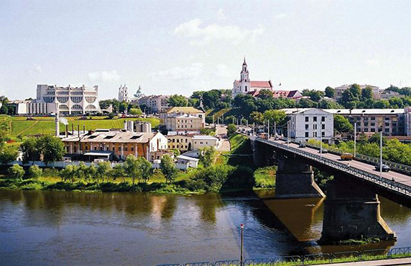 A cityscape with a river, bridge, buildings, a church spire, and lush greenery, suggesting a warm season.