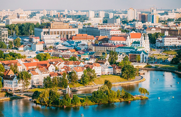 Aerial view of a cityscape with historical buildings, a river meandering through the center, green trees, and open spaces under a clear sky.