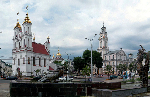 A cityscape with an Orthodox church, a classical building with a clock tower, and a statue under a cloudy sky.