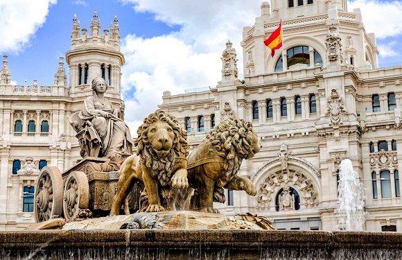 A statue of a figure on horseback accompanied by two lions in front of an ornate building with a Spanish flag.