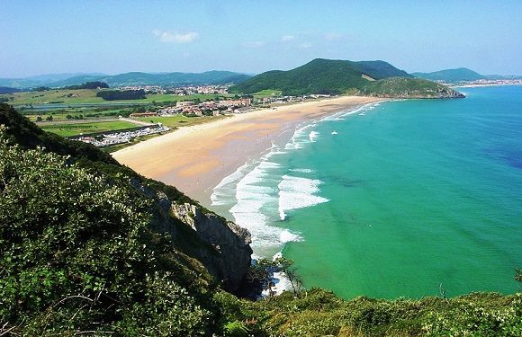 A scenic coastal landscape with a sandy beach bordered by green hills and clear blue waters.