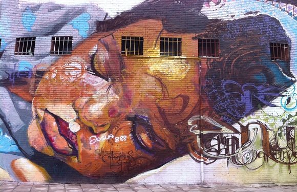 A colorful mural of a person's face in profile view, with abstract elements and graffiti-style lettering on a wall with windows.