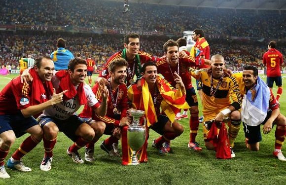 Soccer players in red and yellow uniforms celebrate with a trophy on a grassy field, stadium lights behind them.