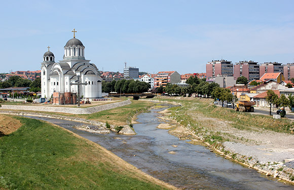 A landscape view of a city with a prominent white church with gray domes near a river, with buildings in the background under a clear sky.