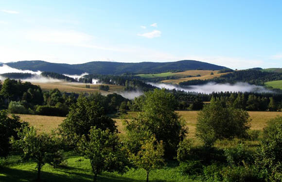 A scenic landscape with rolling hills, patches of forests, and low-lying clouds or mist above the fields under a blue sky with scattered clouds.