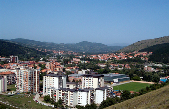 A panoramic view of a town with multi-story residential buildings in the foreground, surrounded by green hills under a clear blue sky.