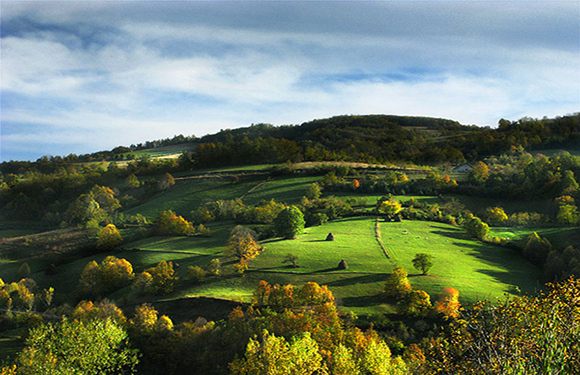 A picturesque landscape with rolling hills, green fields, and scattered trees under a partly cloudy sky.