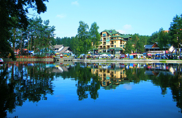 A scenic view of a calm lake reflecting colorful buildings surrounded by trees under a clear blue sky.