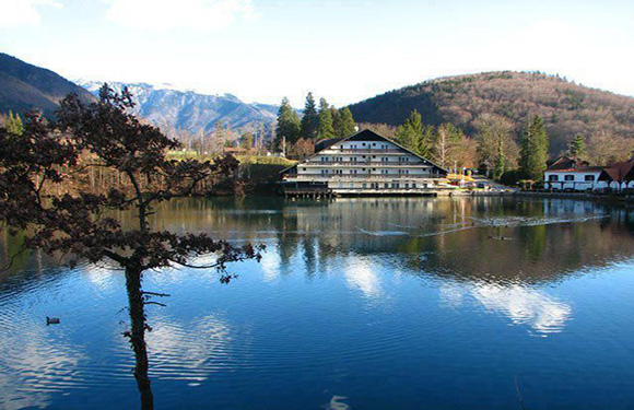 A scenic view of a calm lake reflecting a building and surrounding trees with mountains in the background under a partly cloudy sky.