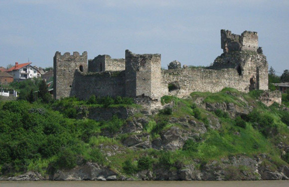 An image of a ruined castle on a hill with green foliage in the foreground and a cloudy sky in the background.