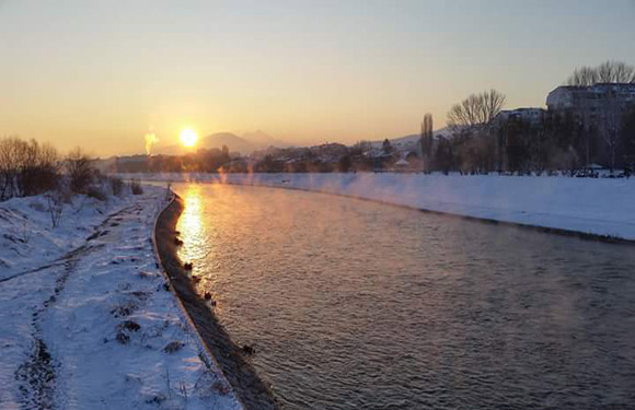 A winter sunset casts a warm glow over a snowy riverbank, calm river, distant trees and buildings under a clear sky.