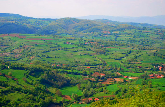 A scenic view of a lush, green hilly landscape with patches of forests and fields, and small buildings scattered throughout.