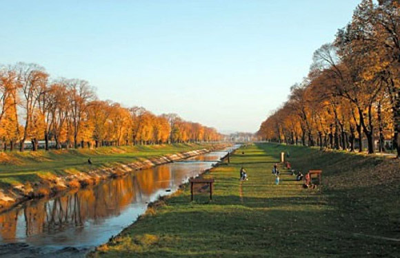 A picturesque canal scene with sky reflections, autumn trees, a grassy path, and distant people enjoying the outdoors.