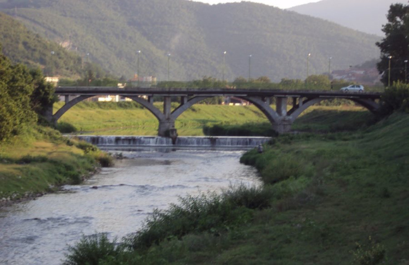 A stone bridge with multiple arches spans over a river, with trees and hills in the background.