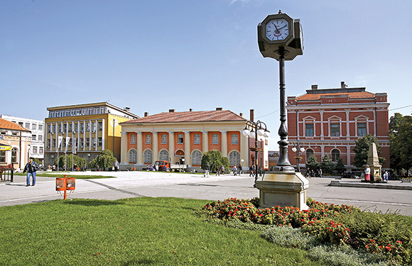 A town square with a clock post in the foreground, flowerbeds, and colorful buildings in the background under a clear sky.