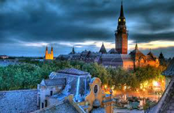 An image of a historic cityscape at dusk with illuminated buildings and a prominent church spire against a cloudy sky.