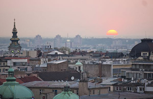 A sunset cityscape features buildings, a large sun, pink-orange sky, and historical domes and a spire.