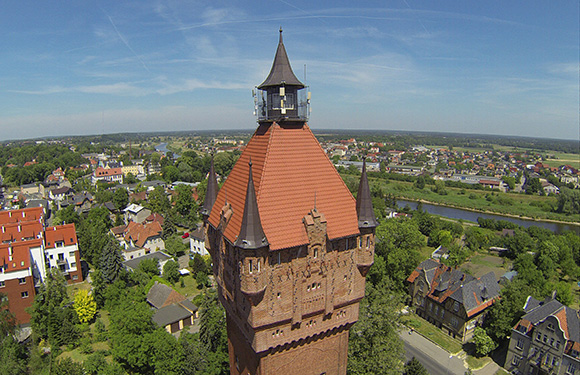 Aerial view of a historic red-roofed tower with a clock, surrounded by green trees and buildings, with a river in the background.