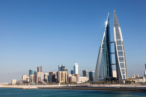 A city skyline with modern skyscrapers, including a prominent twin-towered building, under a clear blue sky, with a coastline in the foreground.