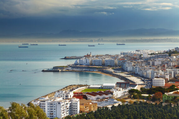 Aerial view of Algiers, the capital of Algeria, showing coastal cityscape with buildings and Mediterranean Sea, under a cloudy sky.