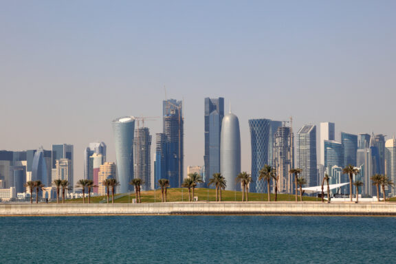 A panoramic view of a modern city skyline with numerous skyscrapers by the waterfront under a clear blue sky.