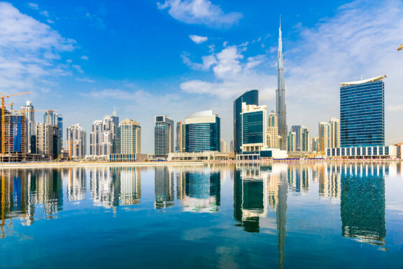 Dubai's skyline, featuring modern skyscrapers and the distant Burj Khalifa, reflected on water under a clear blue sky.