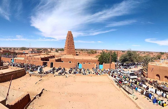 A busy desert market in a town with traditional mud-brick buildings and a tall tower, under a bright blue sky.