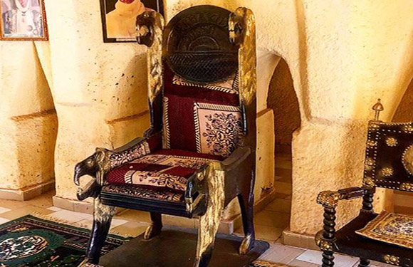 An ornate wooden chair with intricate carvings and a red cushion placed in a room with traditional decor.