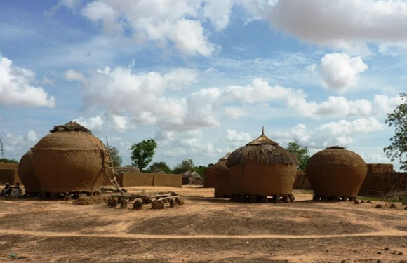 A photo of traditional round mud huts with conical thatched roofs in a rural African setting under a partly cloudy sky.