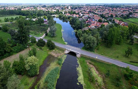 Aerial view of a bridge over a river with adjacent roads, surrounded by greenery and residential areas.