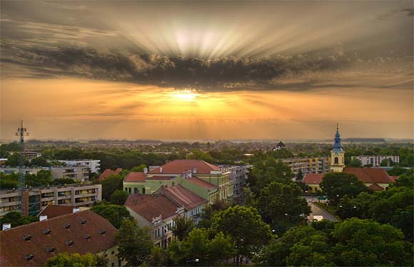 Aerial view of a town at sunset with sun rays piercing through clouds, highlighting buildings and a church spire.