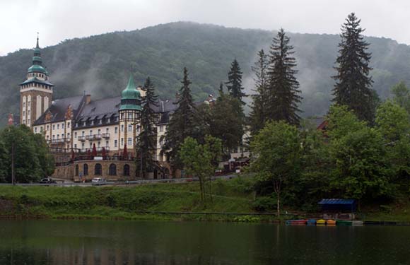 A historic building with towers and spires next to a body of water, with trees in the foreground and misty hills in the background.