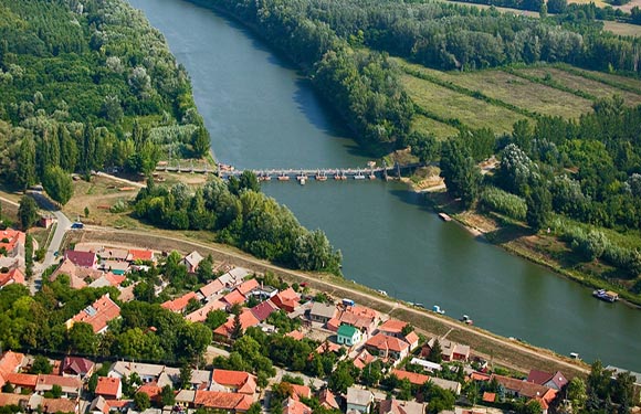 Aerial view of a meandering river with a bridge crossing it, adjacent to a small town with red-roofed buildings surrounded by greenery.