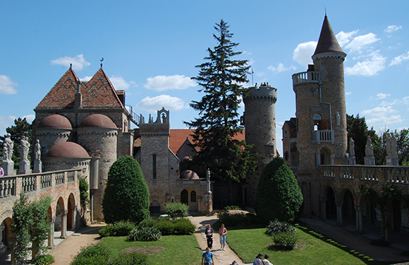 An image of a castle-like structure with multiple towers and pointed roofs, surrounded by a green lawn with a few visitors walking around.