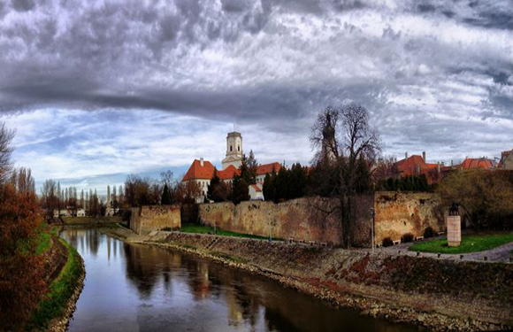 A scenic view of a historic town, featuring a river, stone walls, red-roofed buildings, and a dramatic cloudy sky.