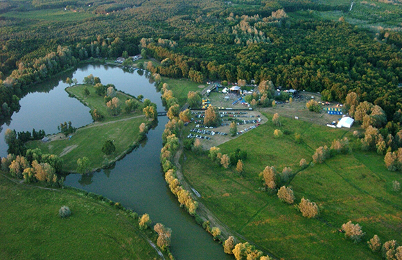 Aerial view of a green landscape with a river, lake, buildings by the water, and trees showing autumn colors.