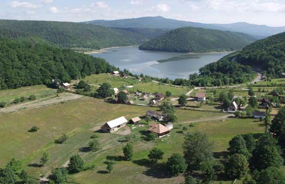 Aerial view of a rural landscape with a village, fields, trees, and a winding river or lake nestled between forested hills.