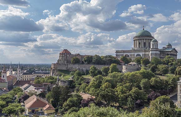 A panoramic view of a historic city with a large domed building and surrounding structures amidst greenery under a partly cloudy sky.