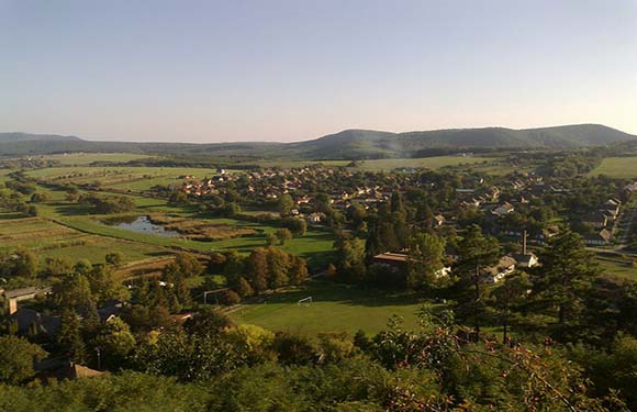 A scenic panorama of a small village amidst green fields, trees, water, and hills under a clear sky.