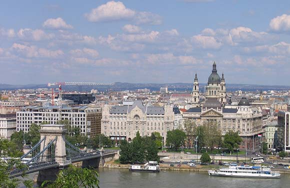 A city panorama featuring historical buildings, a two-spired church, a large bridge, docked boats, and a partly cloudy sky.