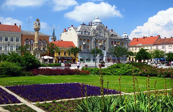 A scenic European town square with colorful buildings, a green park with bright flowers, under a clear blue sky.