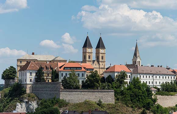 A scenic view of a historic town with prominent twin-spired church, traditional buildings, and greenery under a blue sky with clouds.