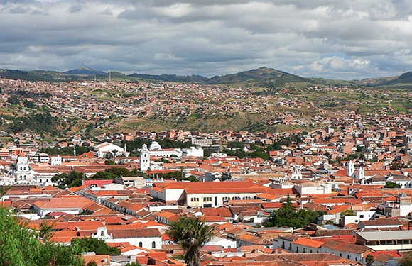 A cityscape with many red-roofed buildings in a valley, surrounded by green hills under a cloudy sky.