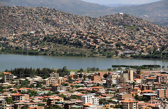 A cityscape with buildings in the foreground near a body of water, with a hill covered in dense buildings in the background under a clear sky.