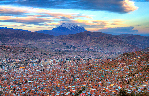 A cityscape panorama of a populous city on hilly terrain, with a snow-capped mountain under a twilight sky.