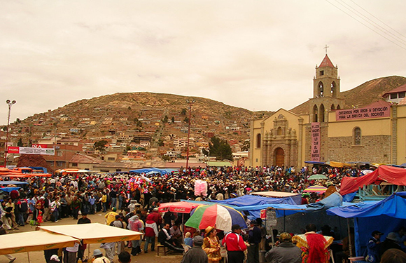 A crowded outdoor market with numerous colorful stalls in front of a church, with a hill in the background.